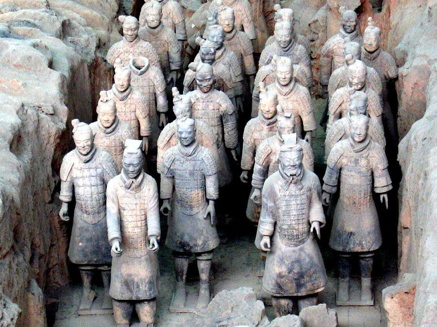 Throughout the entire image, there are life size sculptures of the Terracotta Army of China. These sculptures look very old and dusty, as well as very strong and hardened from the clay they were made out of. There are very strong details on each warrior's armor. Although made of clay, each warrior has their hair tied back with some form of head protection, as well as what appears to be a cloth around their neck. They have a chest of armor and longer skirt like uniforms on to protect their legs, and boots to protect their feet.