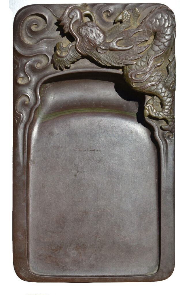 This image shows an example of an inkstone, a square shape with a dark brown color. The majority of the stone has a slope to it to allow the ink to fall and sit at a pool at the bottom of the slope. At the top of this inkstone, there is detailed work of a dragon with his mouth open and almost looks like he is flying in the clouds. This detail work really stands out because of the detail and 3D effect with the carving.